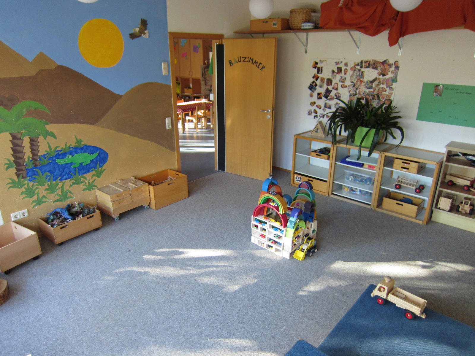 An der rechten Wand steht ein Regal mit Spielsachen, die linke Wand ist mit einer Oasenszene mit Krokodil bemalt. Davor stehen Holzkisten mit Holzklötzen.