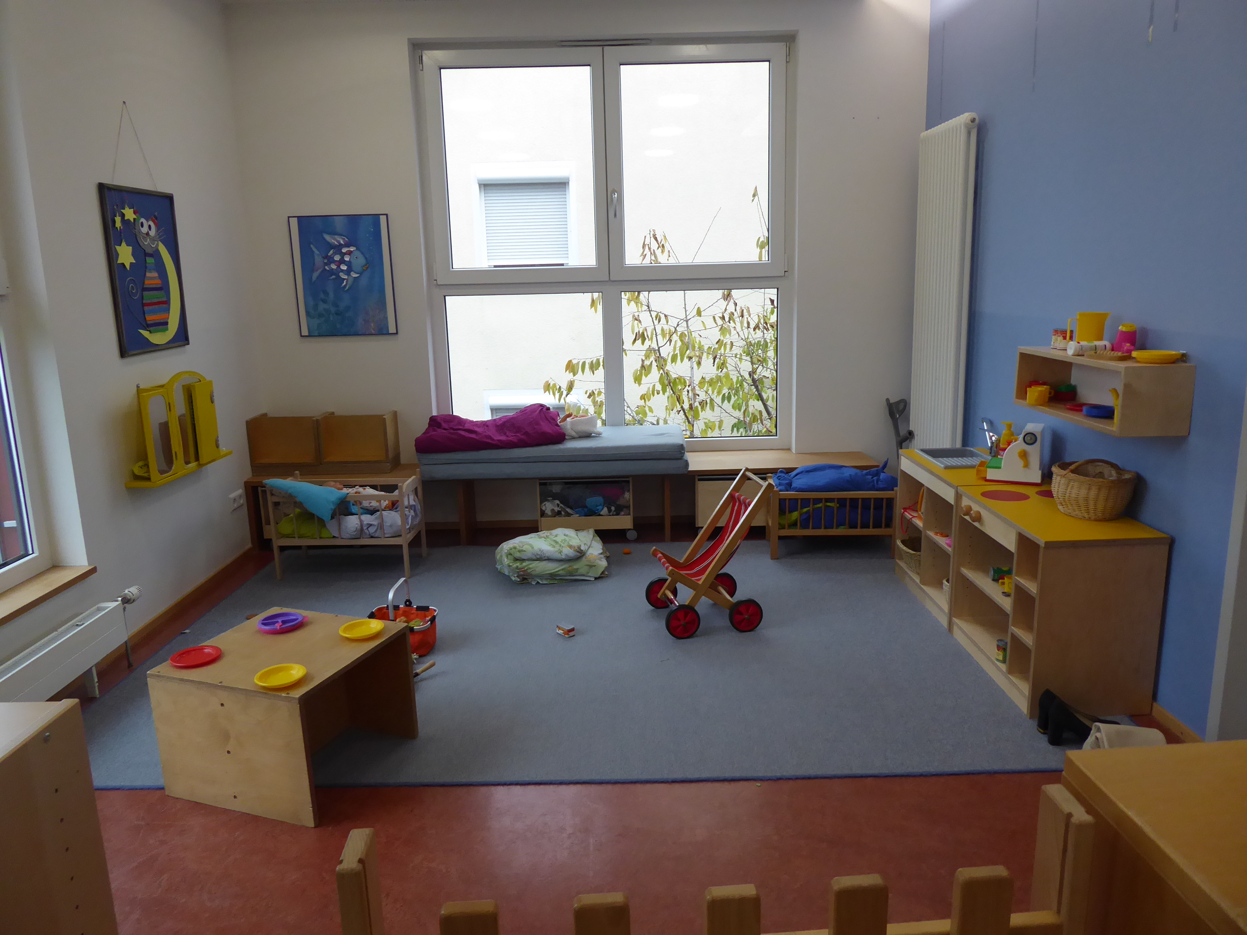 Das Bild zeigt den Rollenspielbereich der Kindergartenkinder. Es gibt zwei Puppenbettchen, einen Kinderwagen und eine Kinderküche.