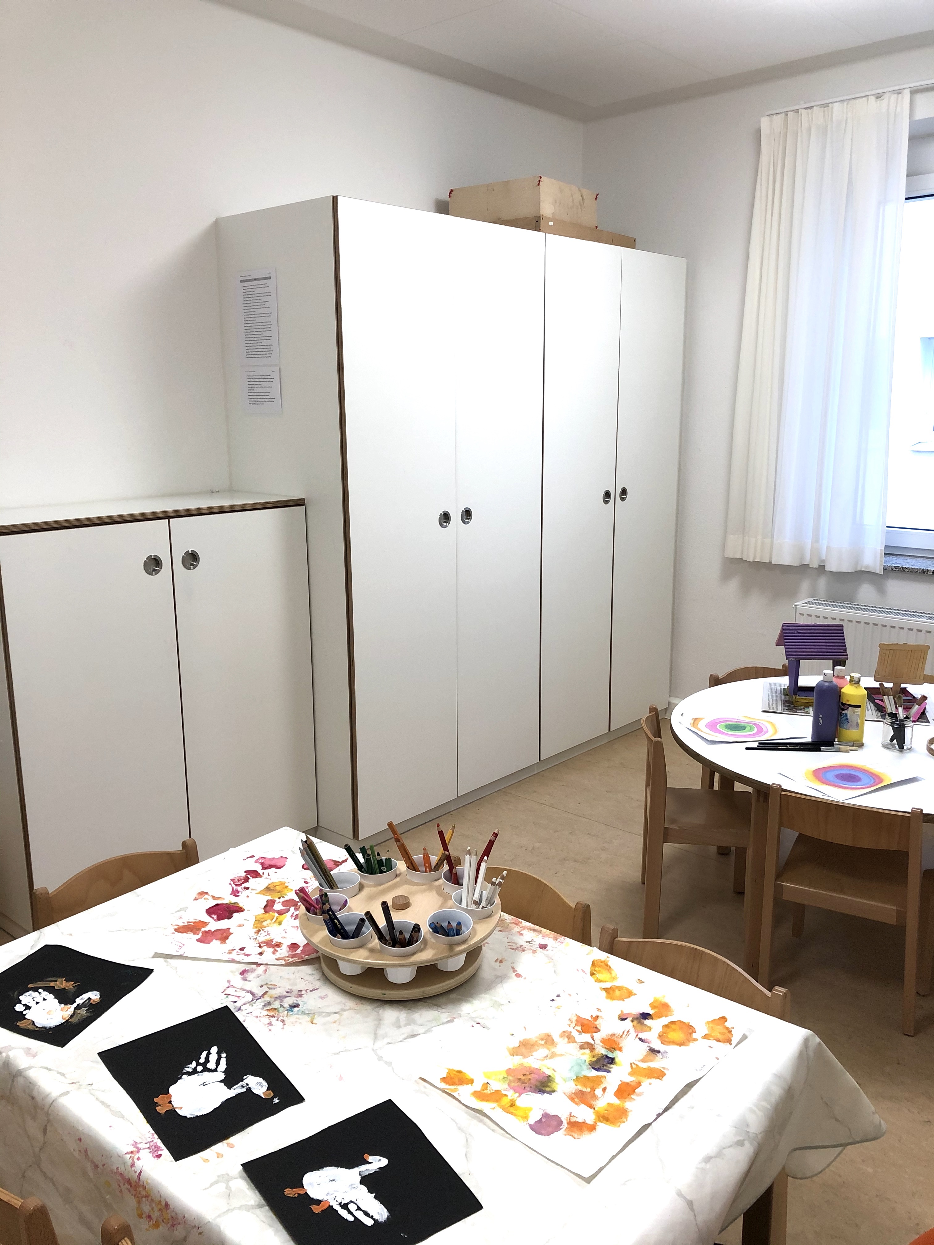 Das Bild zeigt das Atelier im Kindergarten. Es sind drei weiße Schränke an der Wand erkennbar, davor stehen zwei Tische mit Malutensilien.