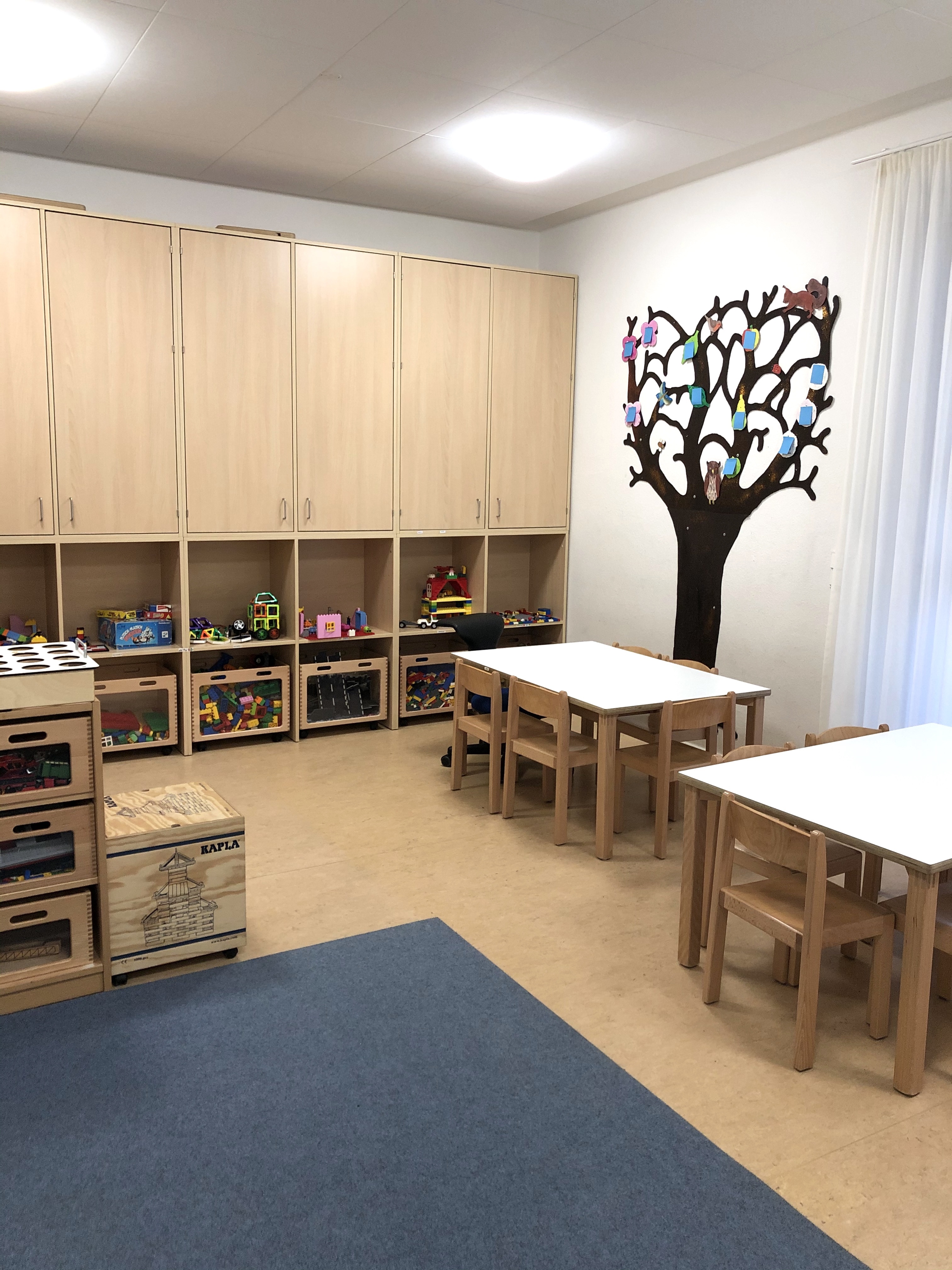 Das Bild zeigt das Bauzimmer im Kindergarten. An der Wand sind Schränke und Regale mit Spielsachen erkennbar. Es sind außerdem zwei Tische mit Stühlen zu sehen.