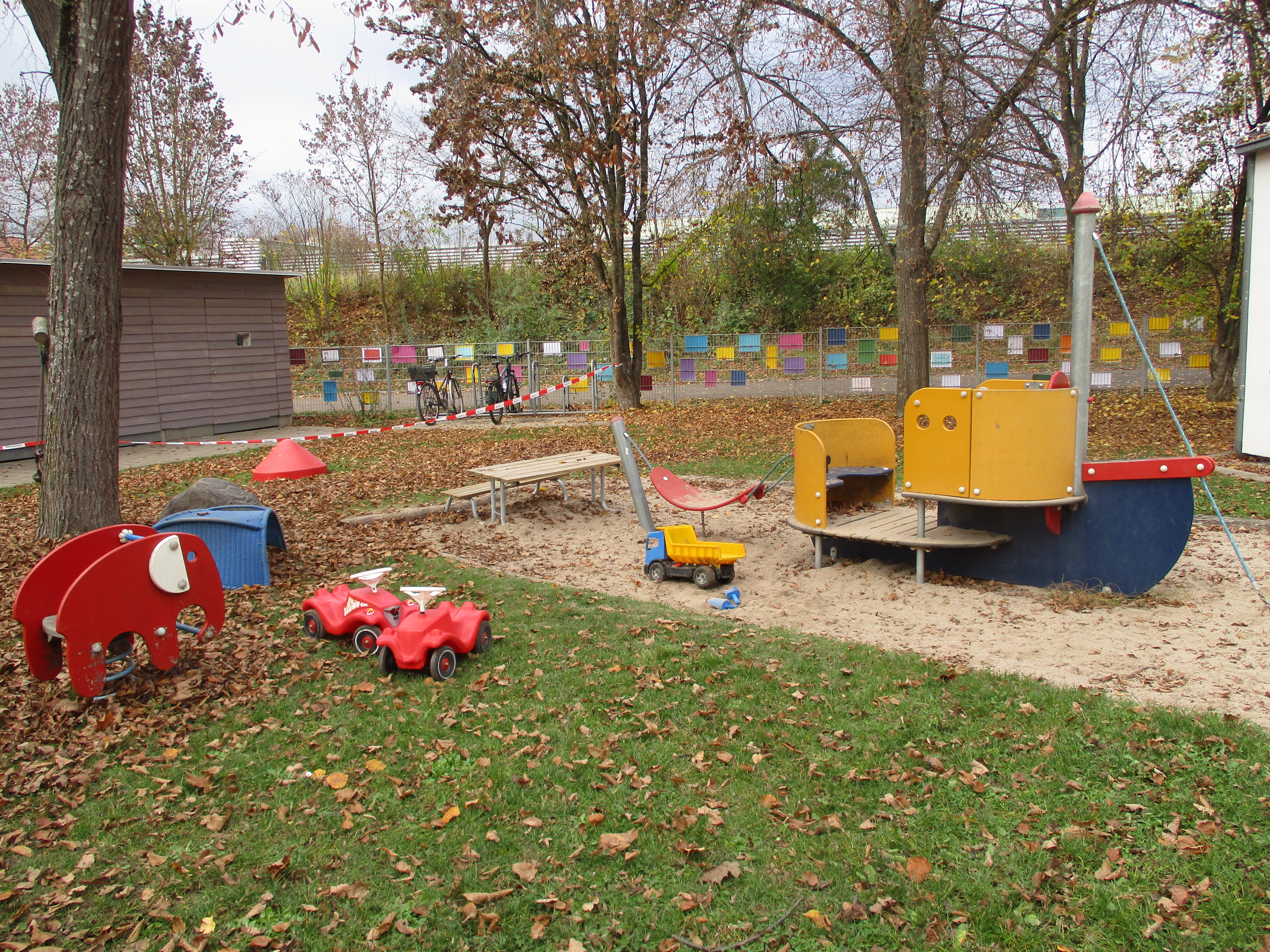 Das Bild zeigt den vorderen Garten des Kinderhaus Feuerhägle. Es ist ein Sandkasten mit einem Spielboot, ein Lastwagen, 2 Bobby Car und ein Schaukelelefant erkennbar.