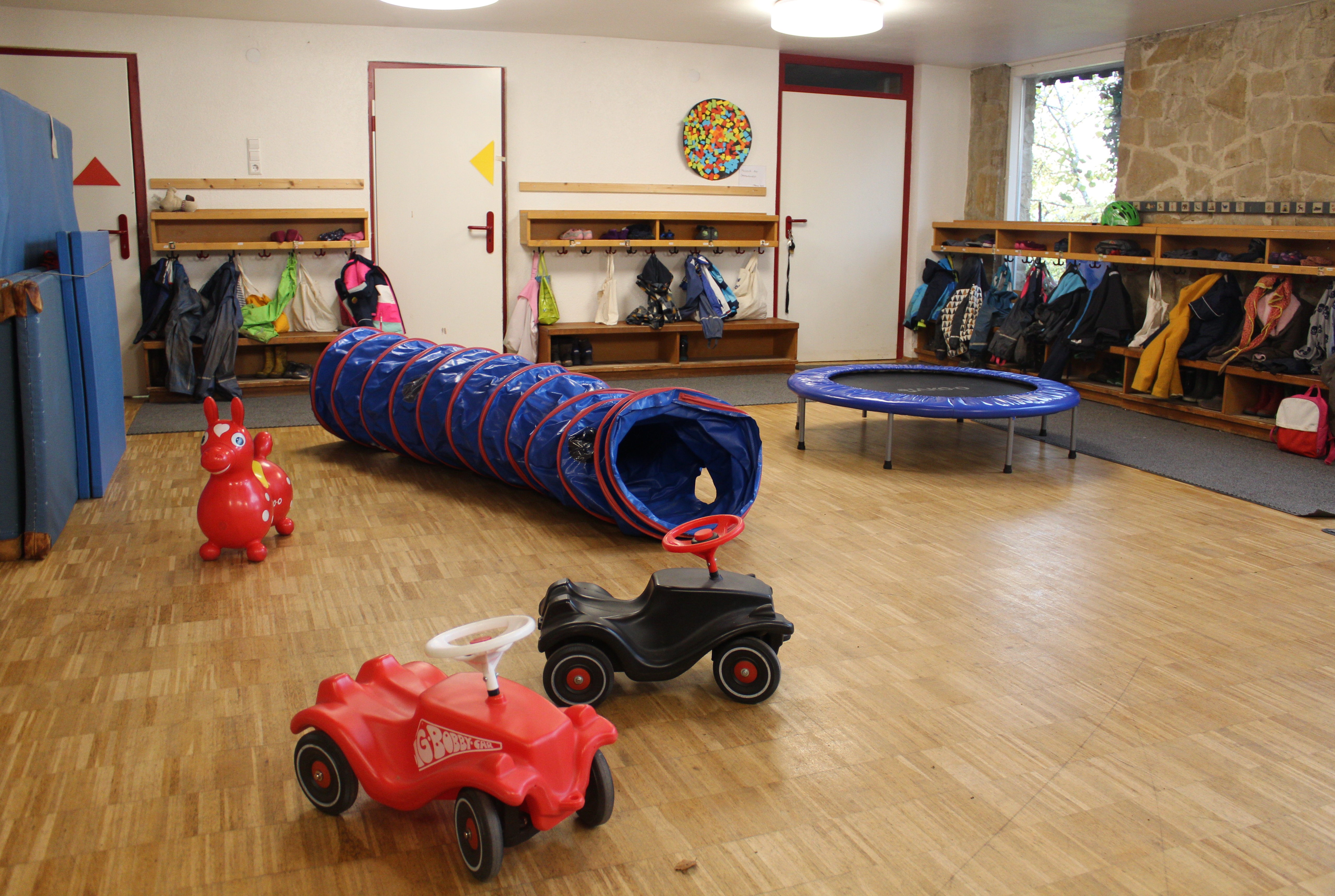 Auf dem Bild sind an den Wänden die Kindergarderoben angebracht, in der Mitte stehen zwei Bobby Cars, ein Trampolin und eine Tunnelröhre.