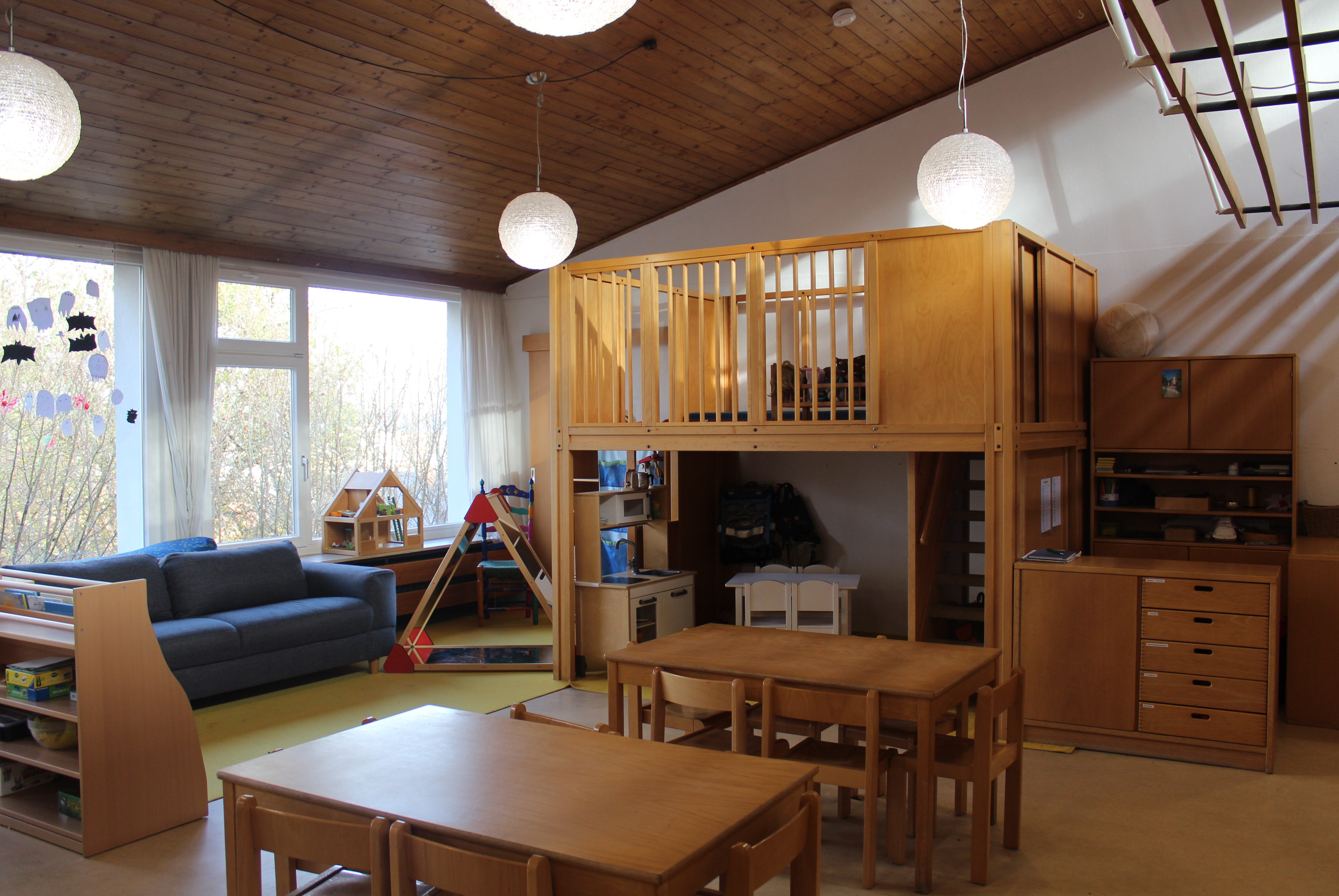 Auf dem Bild ist links am Fenster ein blaues Sofa zu sehen, Eine Kinderküche mit Kindertisch steht im unteren Bereich eines zweistöckigen Spielhauses. Davor stehen 2 Tische mit Stühlen.