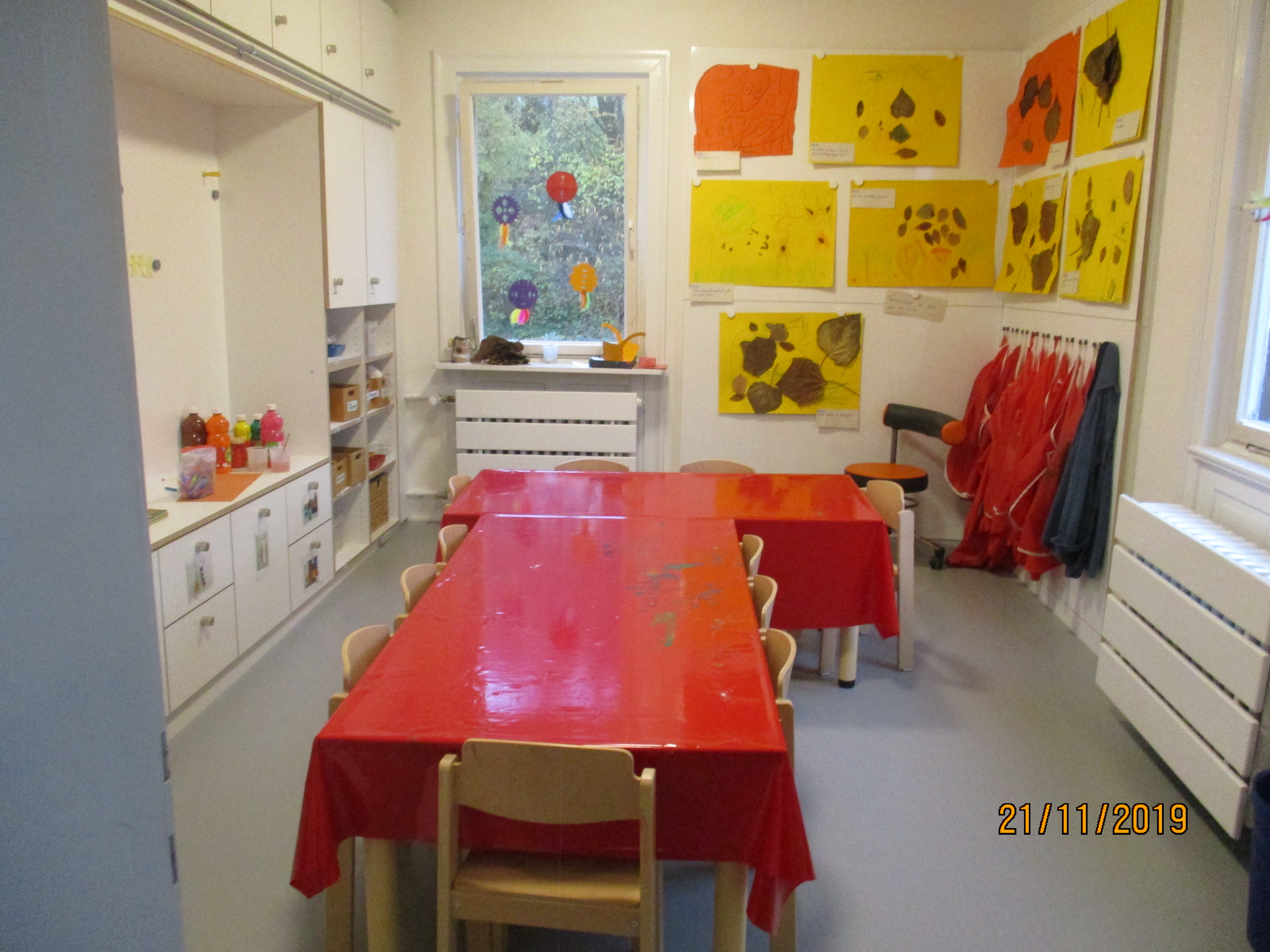 Links sind weiße Einbauschränke mit Farben zu sehen. In der Mitte stehen zwei Tische. Rechts an der Wand hängen Bilder mit Herbstblättern, darunter hängen Malkittel.