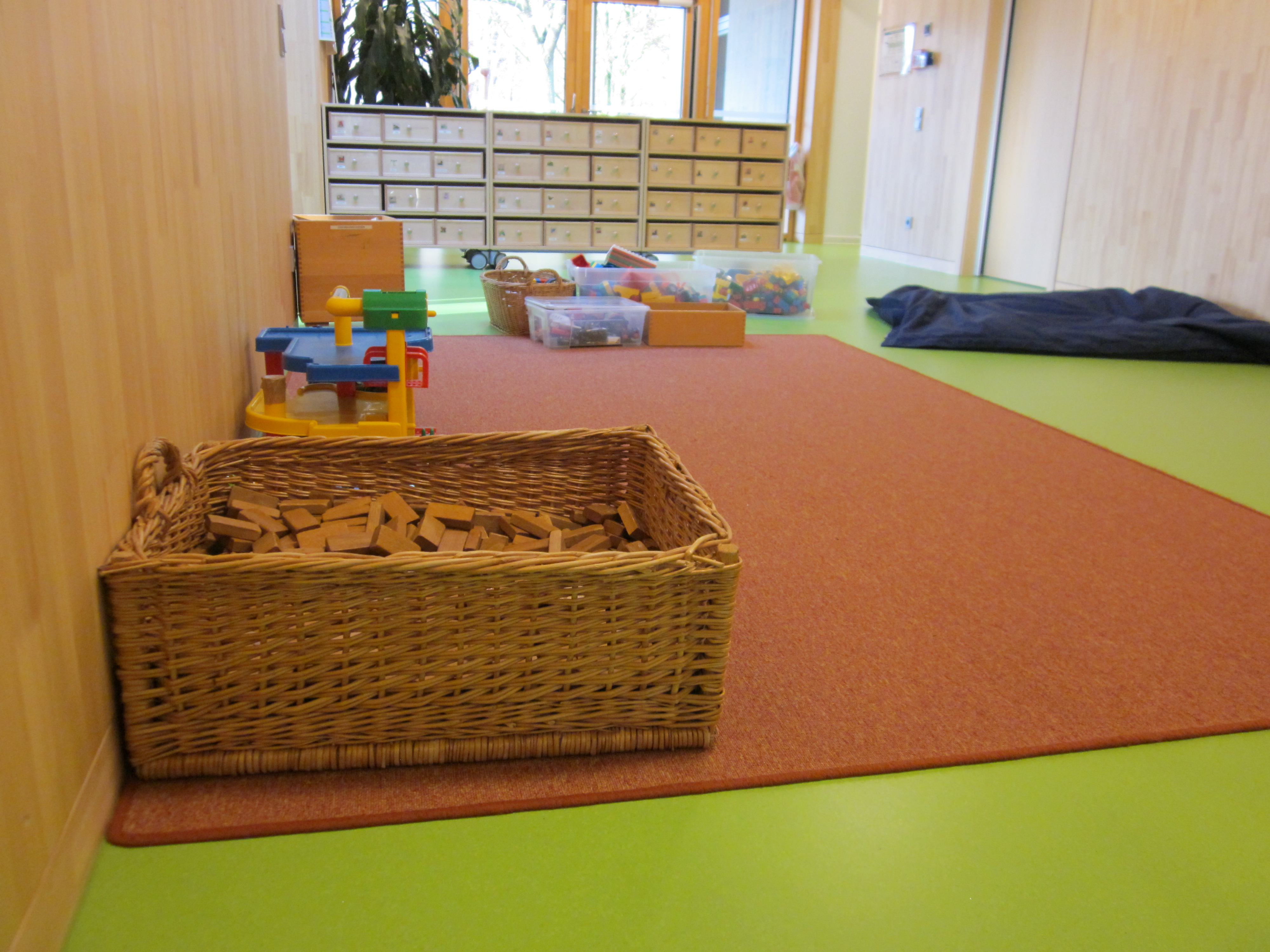 Hier ist die Bauecke im Kindergarten zu sehen. Im Vordergrund steht ein Weidenkorb mit Holzklötzen, dahinter ein Parkhaus und im Hintergrund stehen Boxen mit Bausteinen.