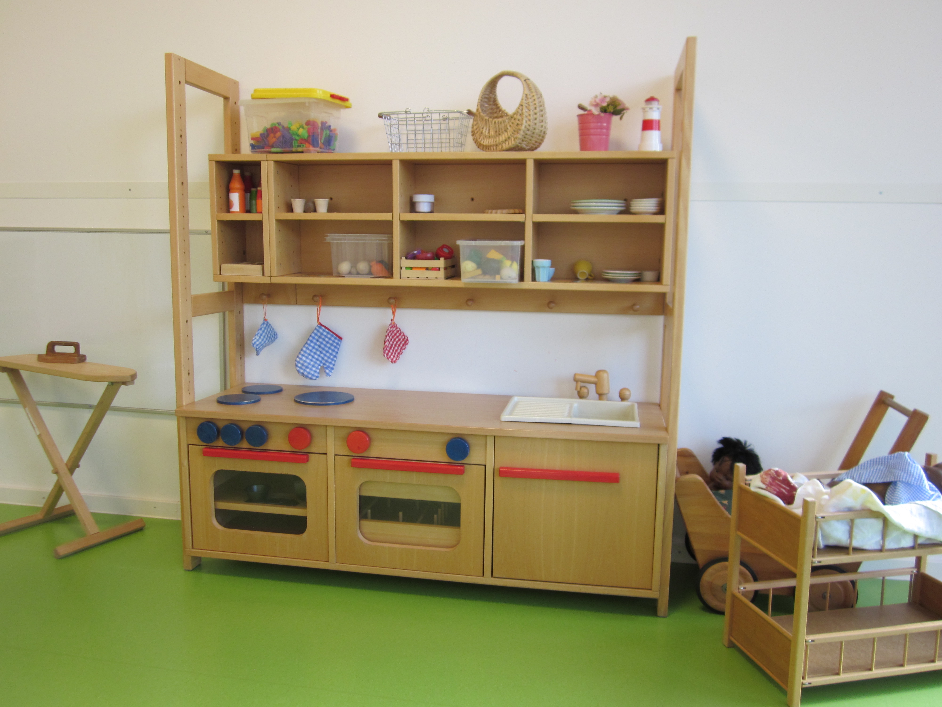 Auf dem Bild ist eine große Kinderküche aus Holz zu sehen. Links daneben steht ein Bügelbrett aus Holz, rechts ein zweistöckiges Puppenbett und ein Puppenwagen.