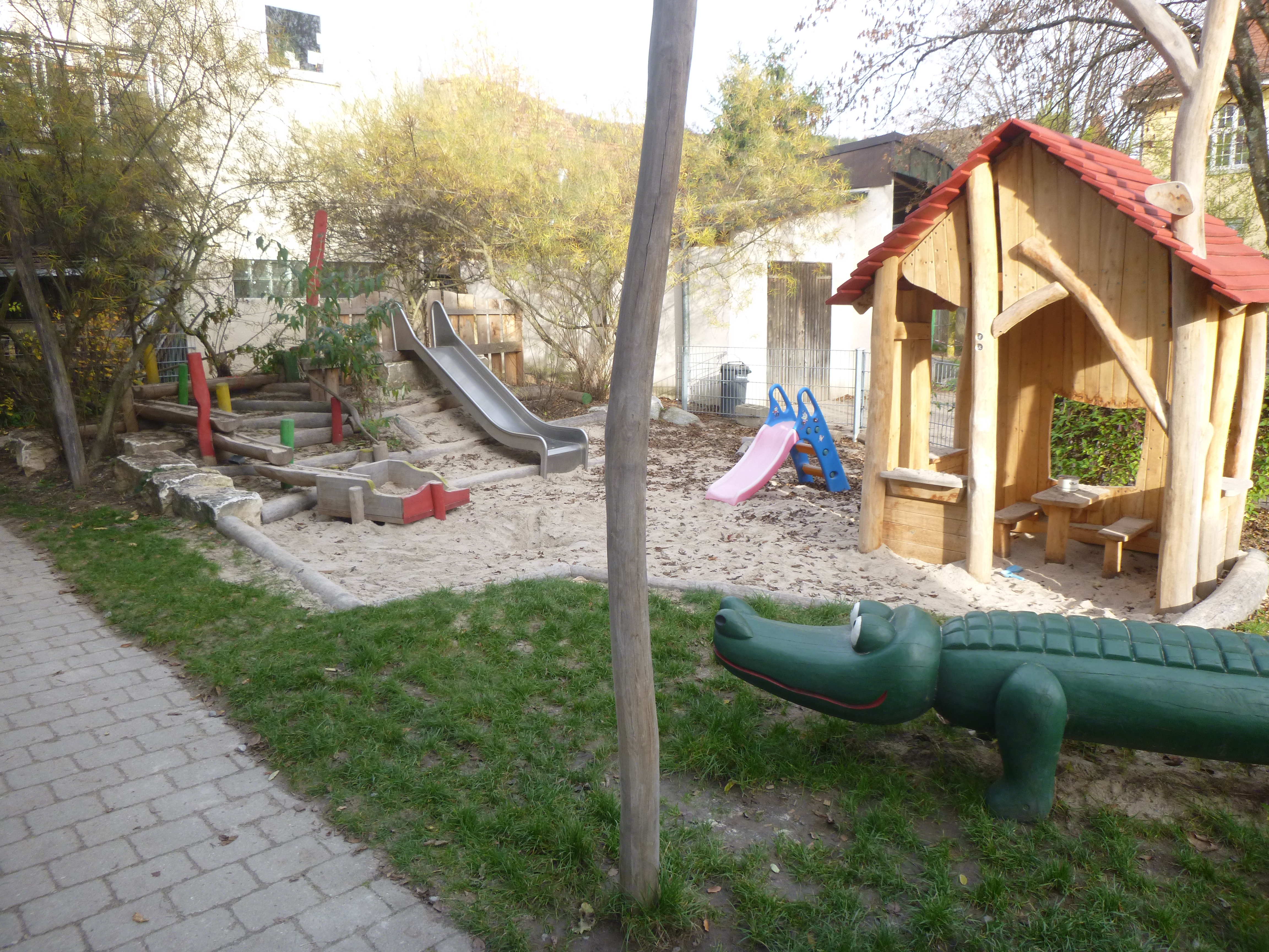 Auf dem Bild ist der Garten der Krippenkinder zu sehen. Rechts im Vordergrund ist ein Krokodil aus Holz zu sehen, dahinter ein Spielhaus aus Holz. Im Sandkasten gibt es außerdem noch zwei kleine Rutschen.