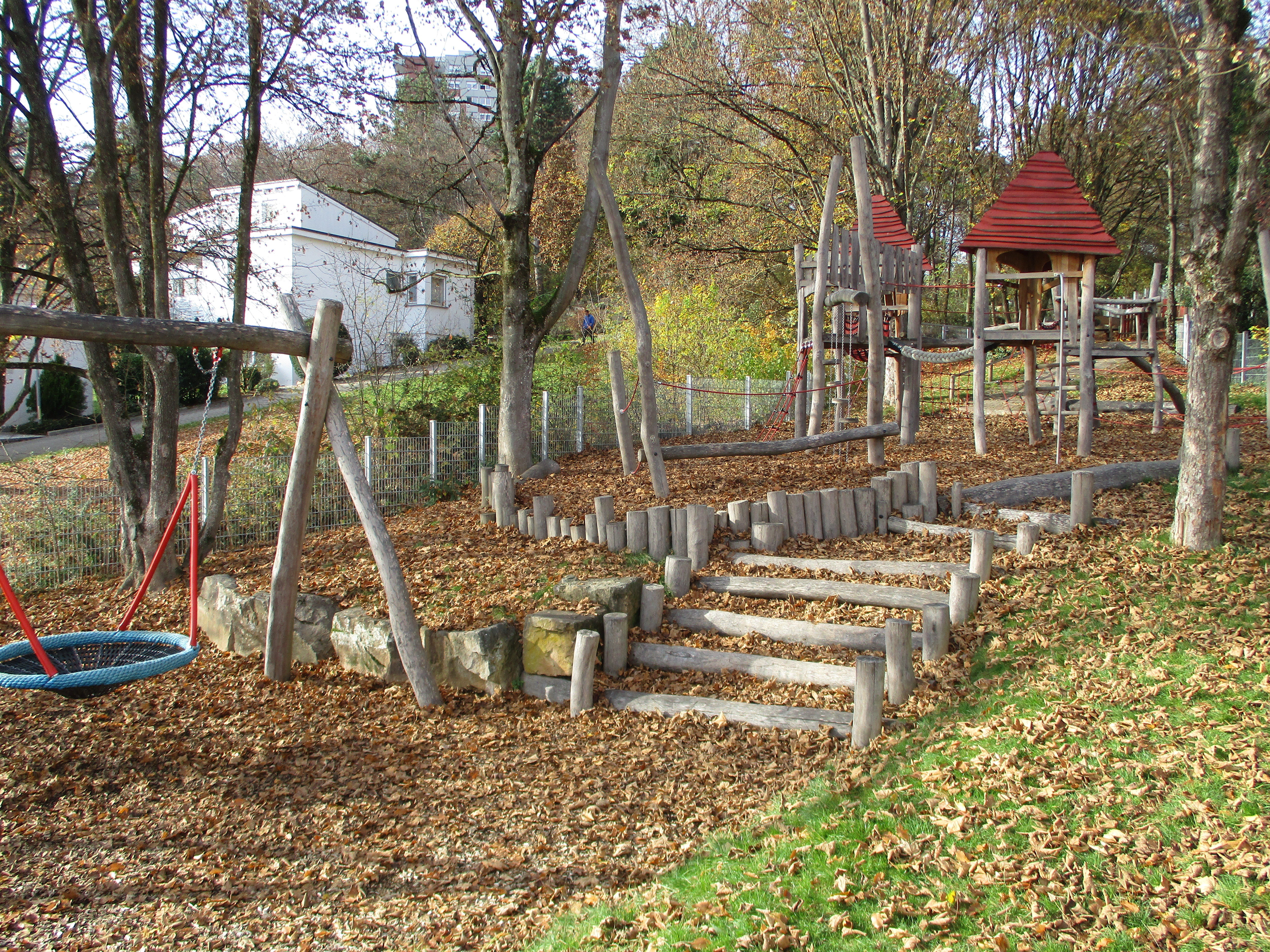 Im Vordergrund ist links eine Nestschaukel, rechts führen Stufen aus Rundholz zu einem Spielgerät aus Holz mit zwei Häuschen zum balancieren und klettern