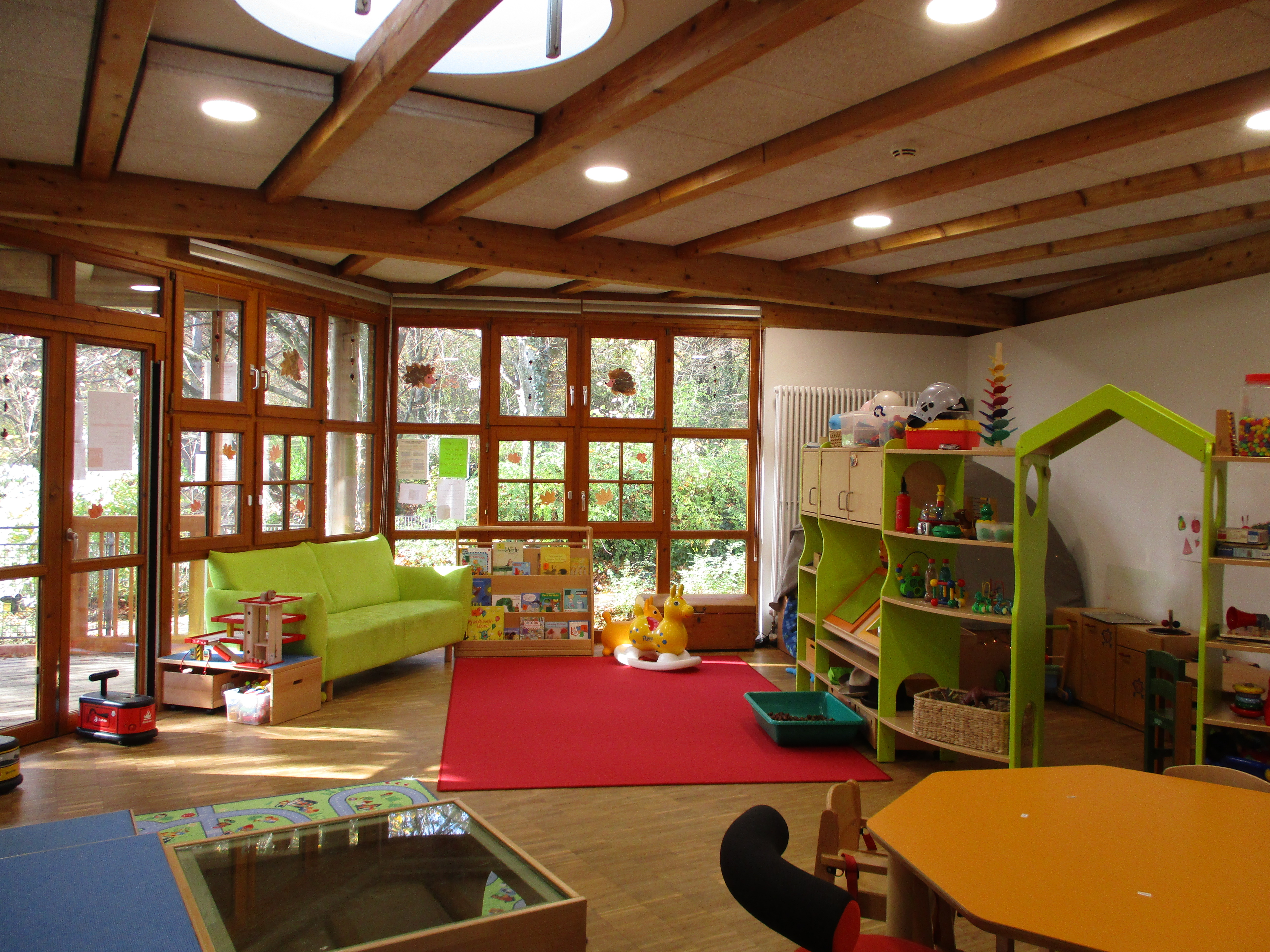 Der Krippenraum ist unterteilt in eine Leseecke mit grünem Sofa, mittig eine Freifläche zum Spielen und hinter einem grünem Regal ist die Rollenspielecke mit Kinderküche.