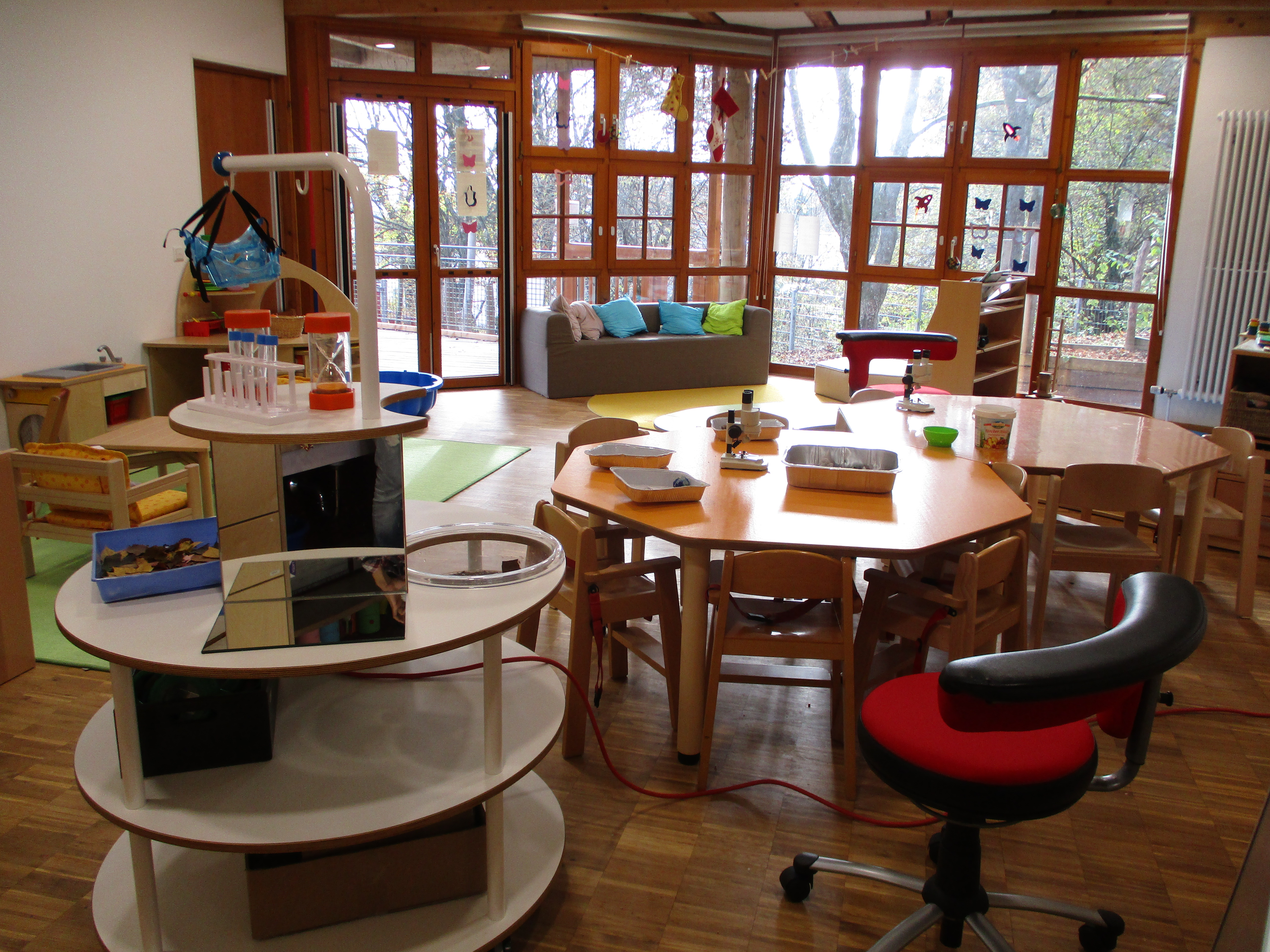 Der Gruppenraum ist unterteilt in eine Leseecke am Fensert mit grauem Sofa mit Kissen, links eine Rollenspielecke. Im Vordergrund steht ein runder Tisch zum forschen, dahinter stehen zwei runde Tische mit Stühlen.