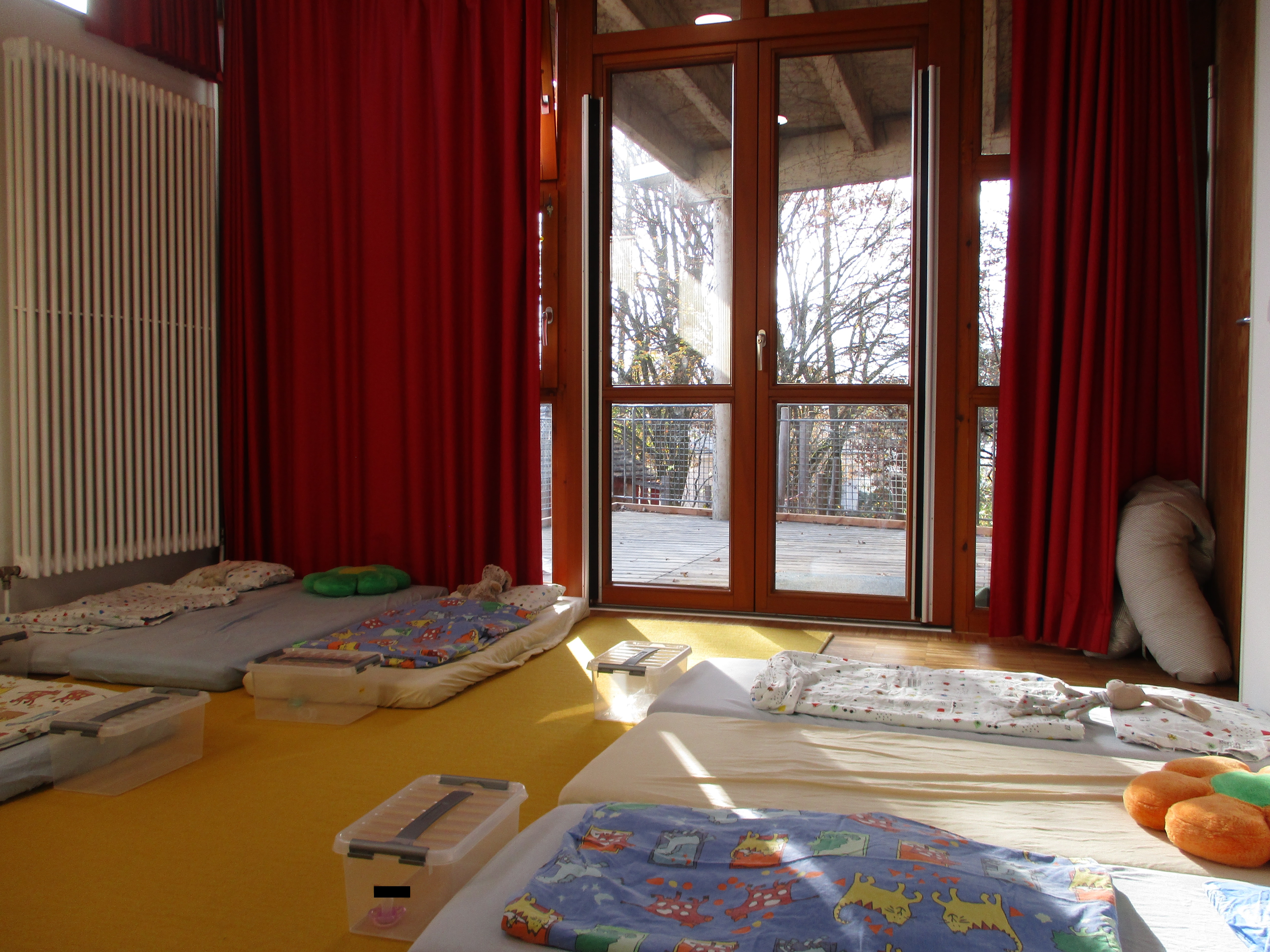 Das Bild zeigt Matratzen mit Bettwäsche auf dem Boden, die Fenster können mit einem roten Vorhang verdunkelt werden.