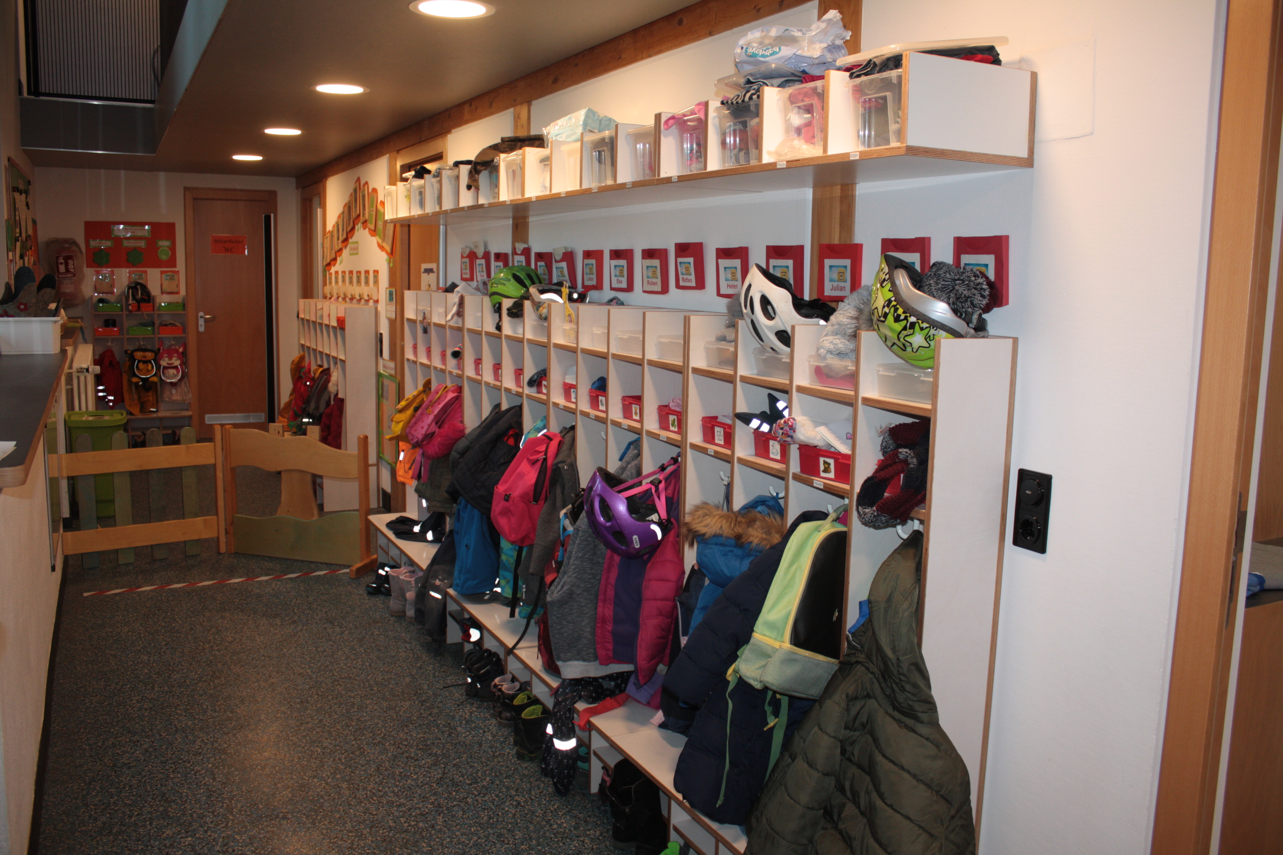 Auf dem Bild ist rechts und in Hintergrund die Garderobe der Kinder. Diese hat verschiedene Fächer für Mützen, Handschuhe und Haken für Jacken.