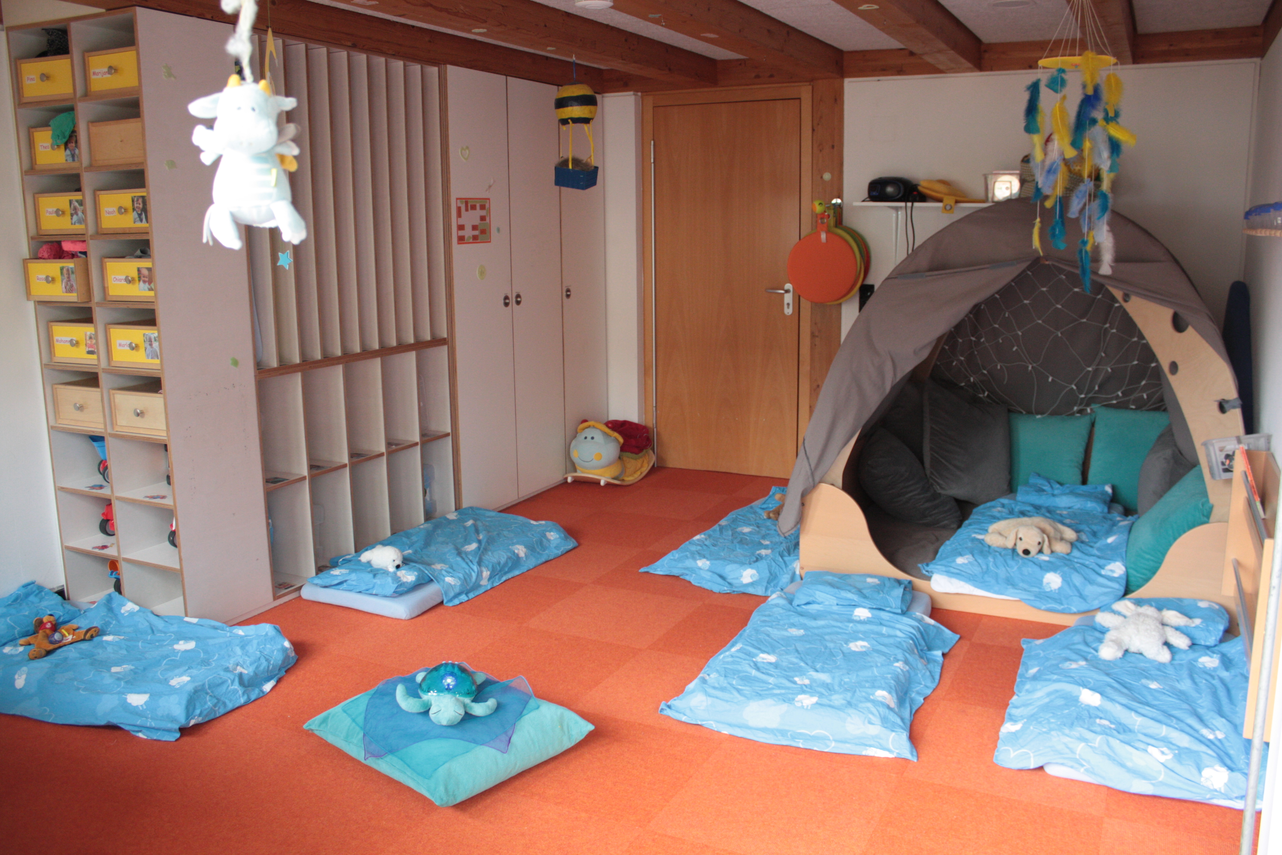 Es sind Matratzen mit blauer Bettwäsche und Kuscheltieren auf dem Boden abgebildet. Links ist ein Einbauregal mit  Schubladen für die einzelnen Kinder und rechts ist eine Kuschelhöhle aus Holz mit vielen Kissen erkennbar.