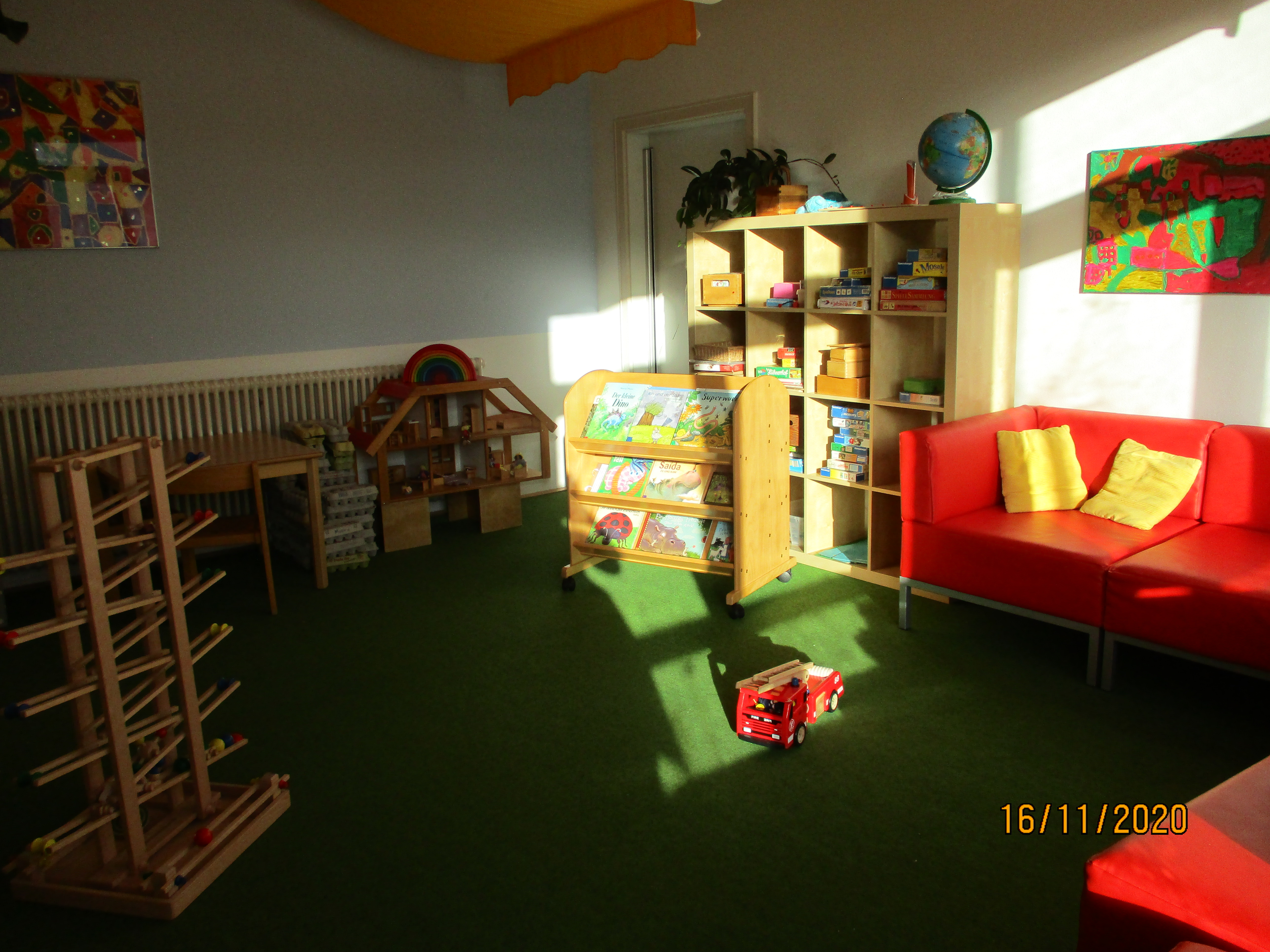 Hier steht an der hinteren Wand ein rotes Sofa mit gelben Kissen. E ist ein Regal mit Büchern und ein weiteres mit Spielen zu sehen. Außerdem ist im Vordergrund eine Kugelbahn ersichtlich.