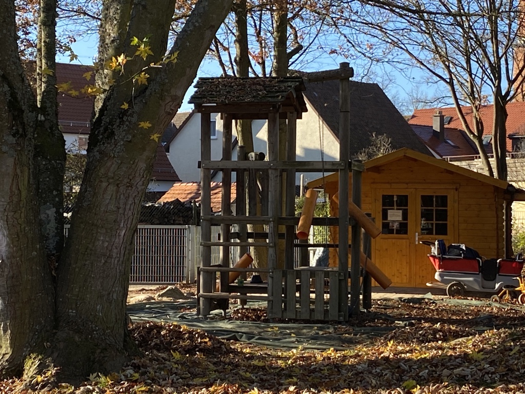 Im Hintergrund ist eine Gartenhütte zu erkennen, in der Bildmitte ist ein Spielhaus aus Holz zu sehen und im Vordergrund ist ein großer Baum abgebildet.