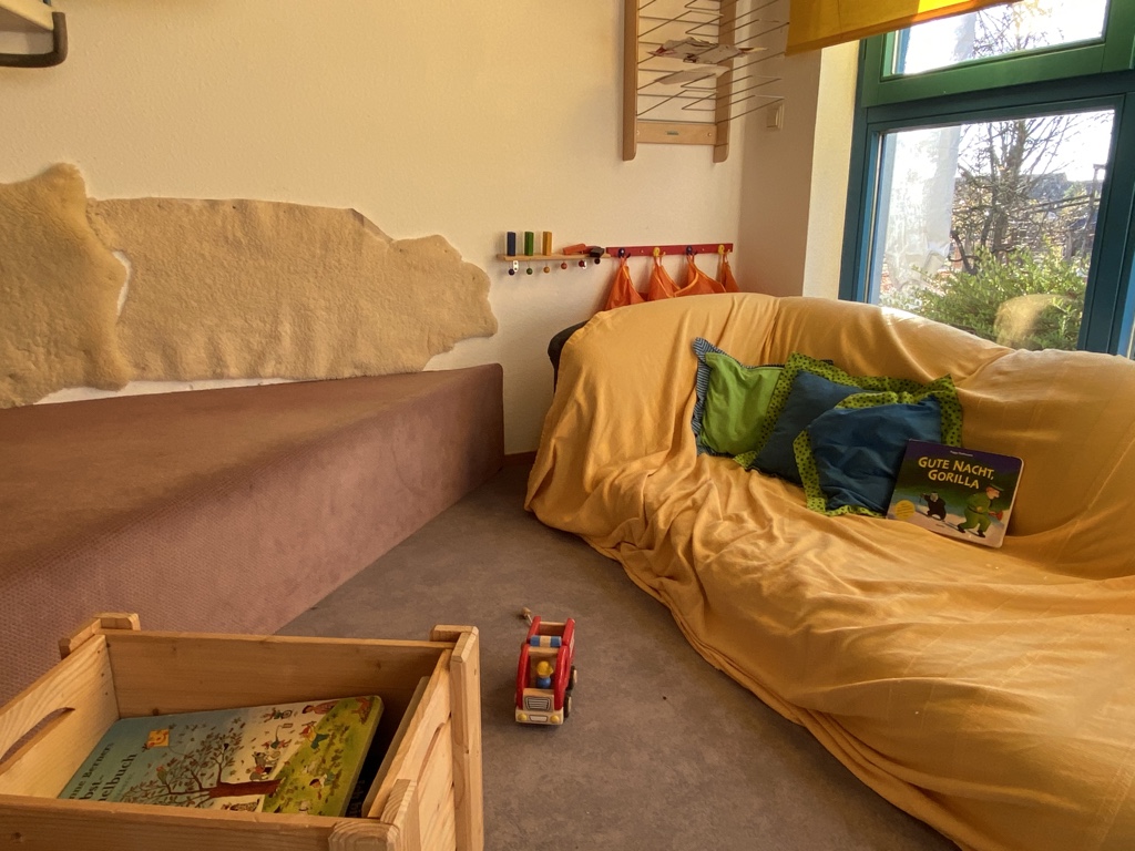 Rechts im Bild steht ein Sofa mit einem gelben Überwurf. Blaugrüne Kissen und ein Buch sind darauf drapiert. Im Vordergrund steht eine Holzkiste mit Büchern. Links ist eine weitere Sitzfläche für die Kinder.