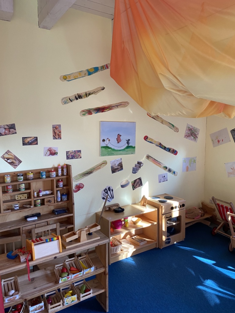 das Bild zeigt einen Kaufladen aus Holz. Rechts daneben ist eine Kinderküche aufgebaut. An der Wand hängen verschiedene Bilder.