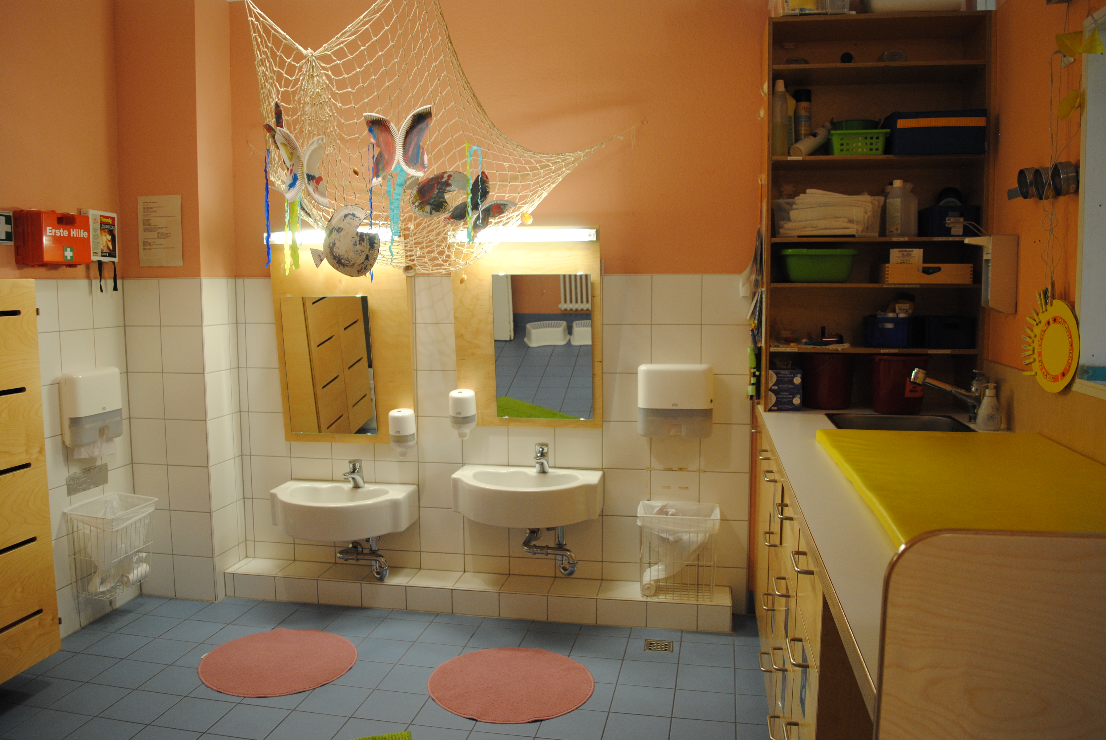 Das Bild zeigt zwei Waschbecken und unterschiedlicher Höhe und Spiegeln. Rechts ist der Wickelbereich. An der Decke wurde ein Netz mit Schmetterlingen als Dekoration aufgehängt.