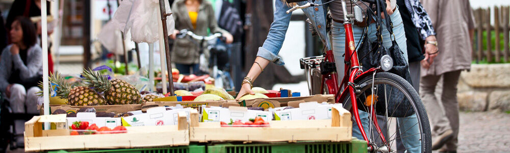 Eine Frau steht mir ihrem Fahrrad an einem Wochenmarktstand und greift nach Obst und Gemüse.