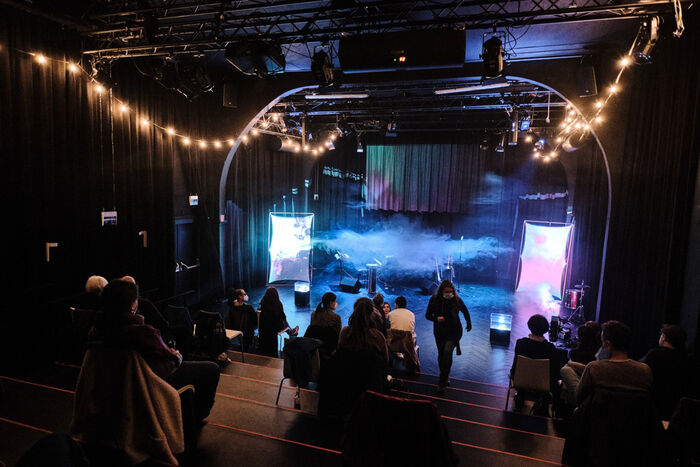 Das Bild zeigt Zuschauer und eine hell erleuchtete Bühne bei einer Vorstellung im Zimmertheater.
